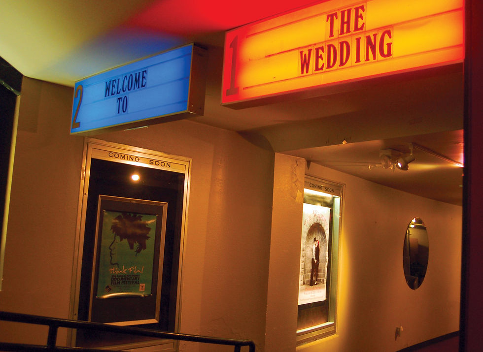 Movie theater entrance with marquee signs "Welcome to" in blue and "The Wedding" in red. Posters below read "Coming Soon."