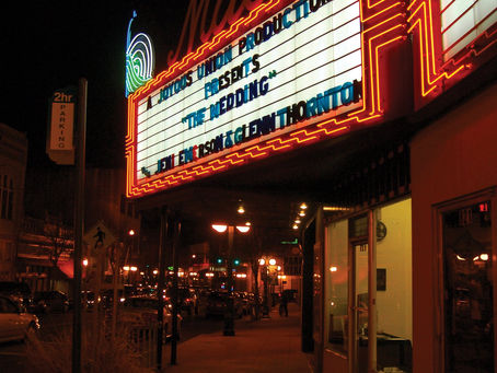 Illuminated theater marquee from the Malco Theatre in Hot Springs, AR at night reading "A Joyous Union Production Presents: The Wedding" Street view of Central Avenue with glowing lampposts and parked cars.