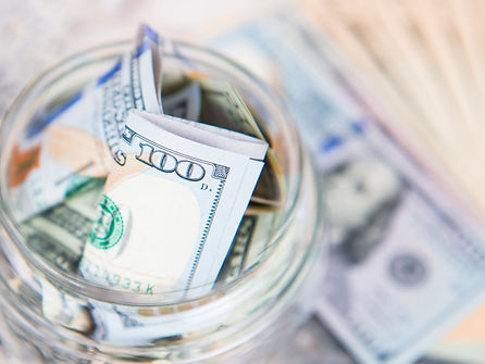 A mason jar full of money with a hundred dollar in US currency on top.
