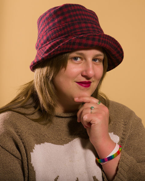 Lea in a hat and sweater ponders on a wheat-coloured background.