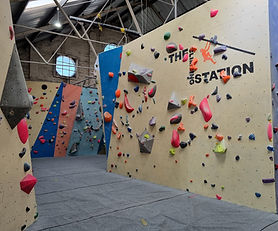 The Climbing Station indoor climbing gym with various bouldering walls, some angled and others vertical, covered in diverse, brightly colored hand and footholds. "THE CLIMBING STATION" is written on the main wall.