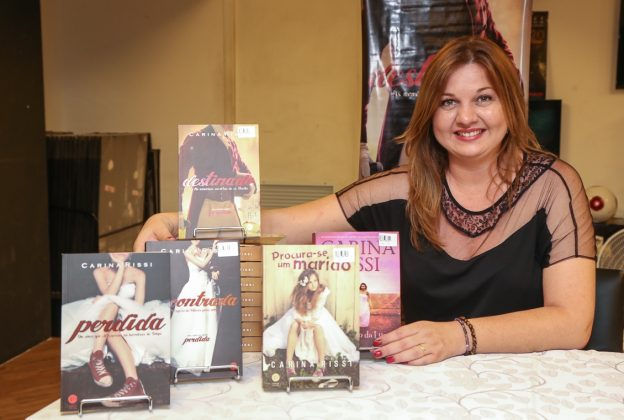 Carina posa sentada e sorrindo apoiada em uma mesa, ao lado dos livros "perdida", "encontrada", "destinada, "procura-se um marido"
