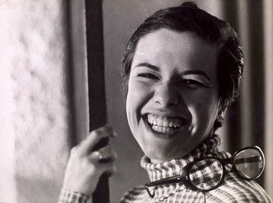 Na imagem: Elis Regina sorri para a câmera, com cabelo curto, um suéter listrado e um óculos pendurado no ombro.