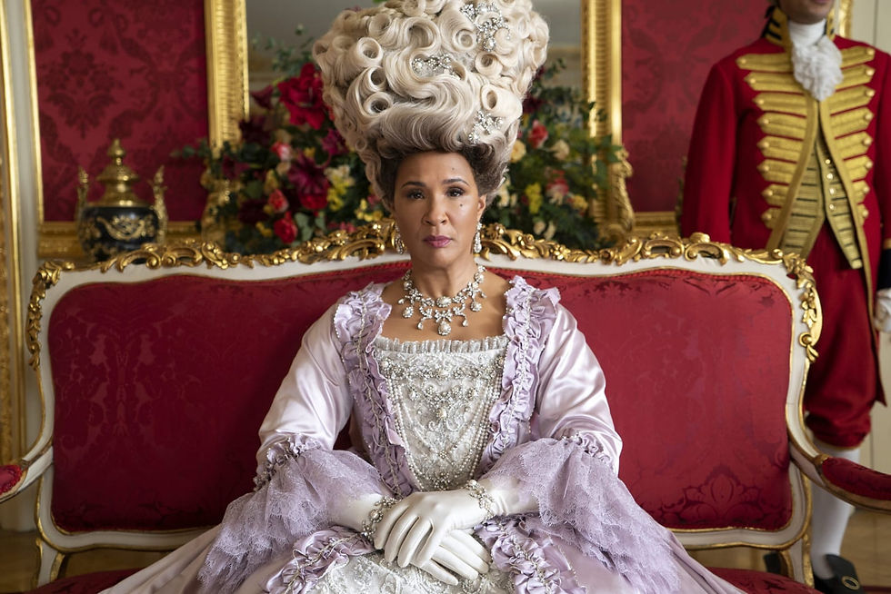 A rainha Charlotte está sentada em um sofá antigo de estofado vermelho, com detalhes em ouro, usando uma peruca enorme branca cheia de cachos e joias, usa também um colar de diamantes e um bonito vestido bufante.