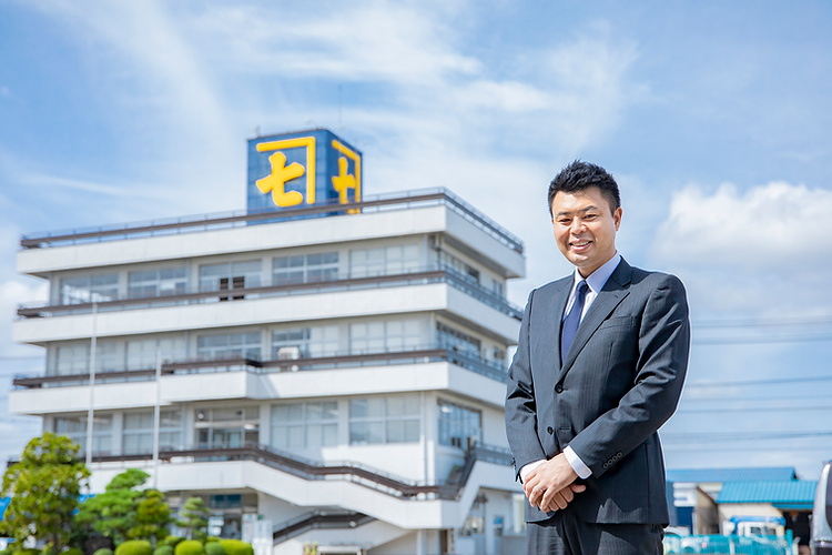 社屋を背に笑顔を見せる河上金物株式会社 代表取締役 河上森の様子