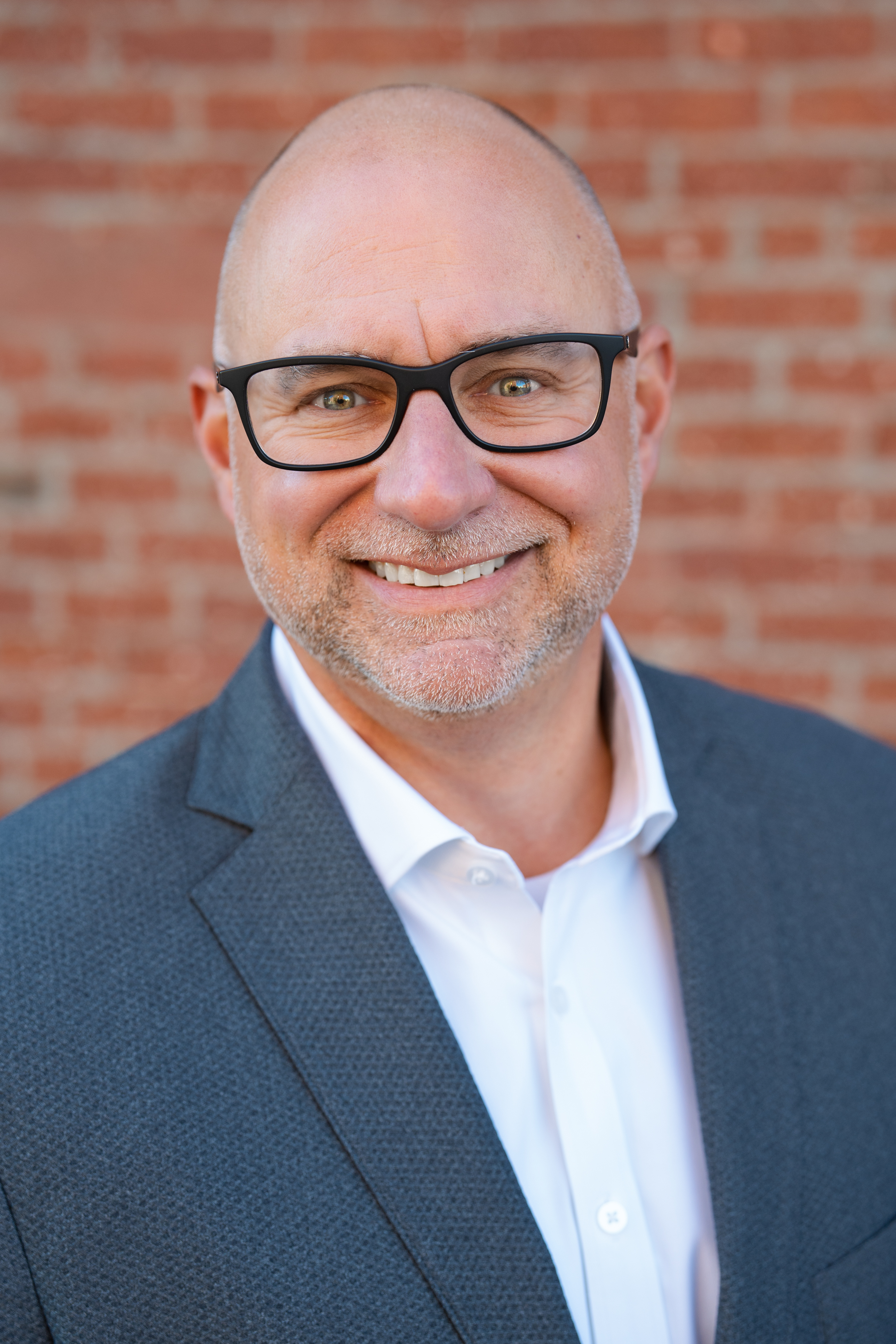 Professional headshot of Dan Girard with dark rimmed glasses with a brick wall behind him