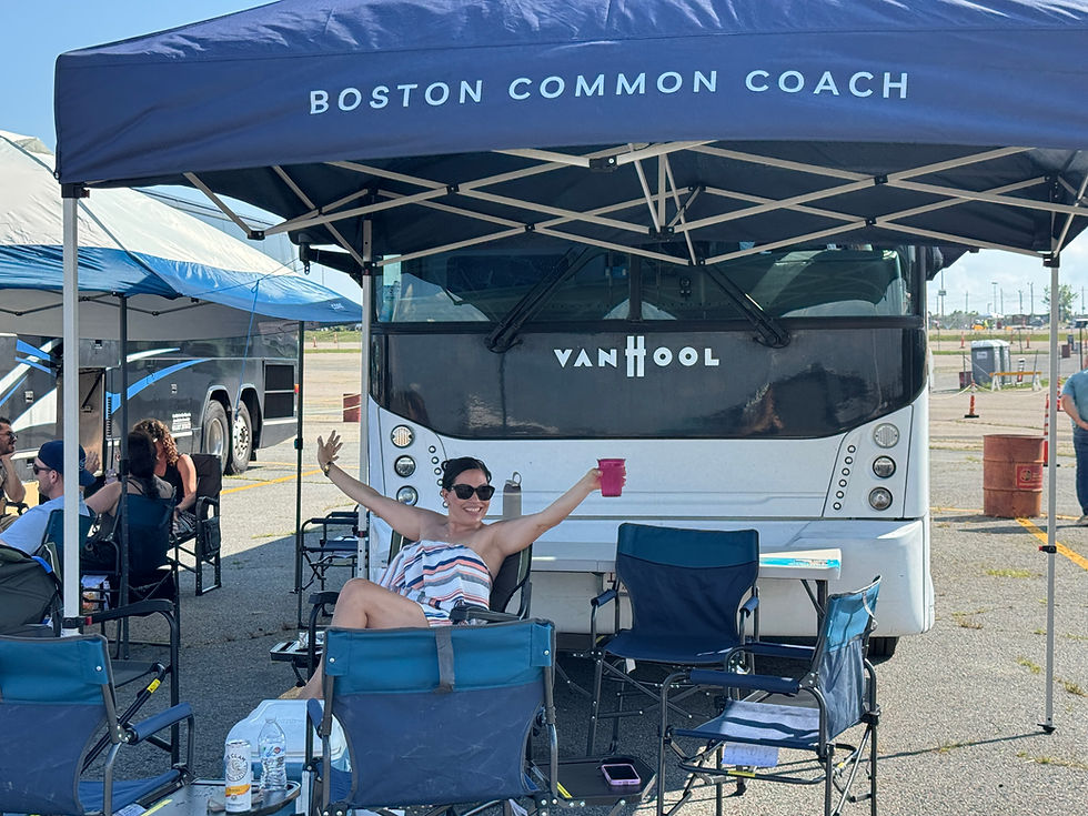 Woman sitting in a camping chair under a Boston Common Coach branded tent in front of a coach bus with arms out with a beverage in hand as part of a tailgate.