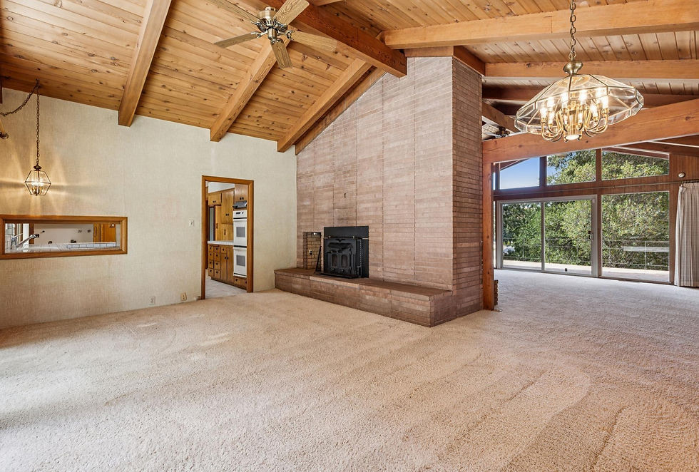 Spacious room with high wooden ceiling, brick fireplace, chandelier, and large windows overlooking greenery. Neutral tones set a cozy mood.