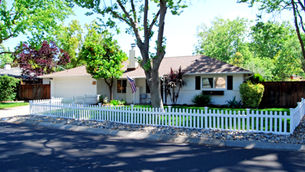 Single-story house with white fence, green lawn, and trees. American flag on porch. Bright day, peaceful suburban setting.