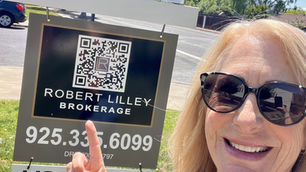 Smiling woman in sunglasses points to "Open Tuesday 11-1pm" real estate sign. Green grass and trees in the sunny background.