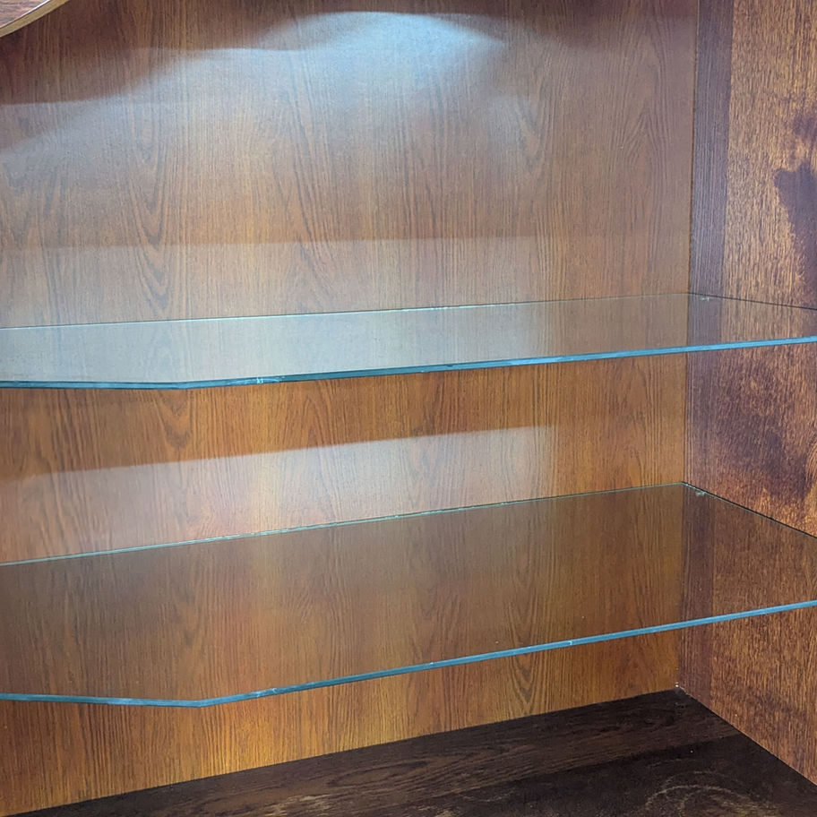 Klasikinė rudo medžio indauja su stiklinėmis durimis ir stalčiais, pastatyta prieš pilką foną šalia odinio krėslo.