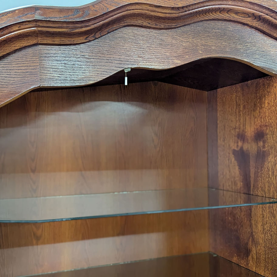 Klasikinė rudo medžio indauja su stiklinėmis durimis ir stalčiais, pastatyta prieš pilką foną šalia odinio krėslo.