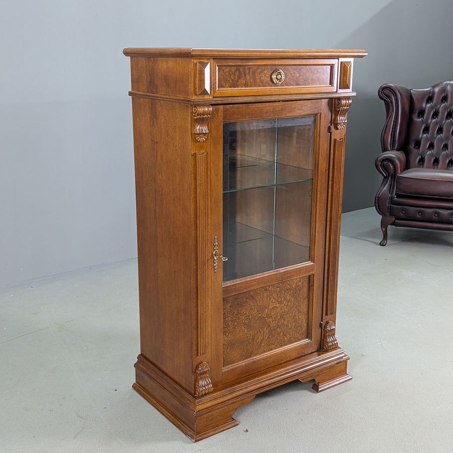 Klasikinė vitrina su stiklinėmis lentynomis ir stalčiumi, pastatyta prieš pilką foną šalia odinio krėslo.