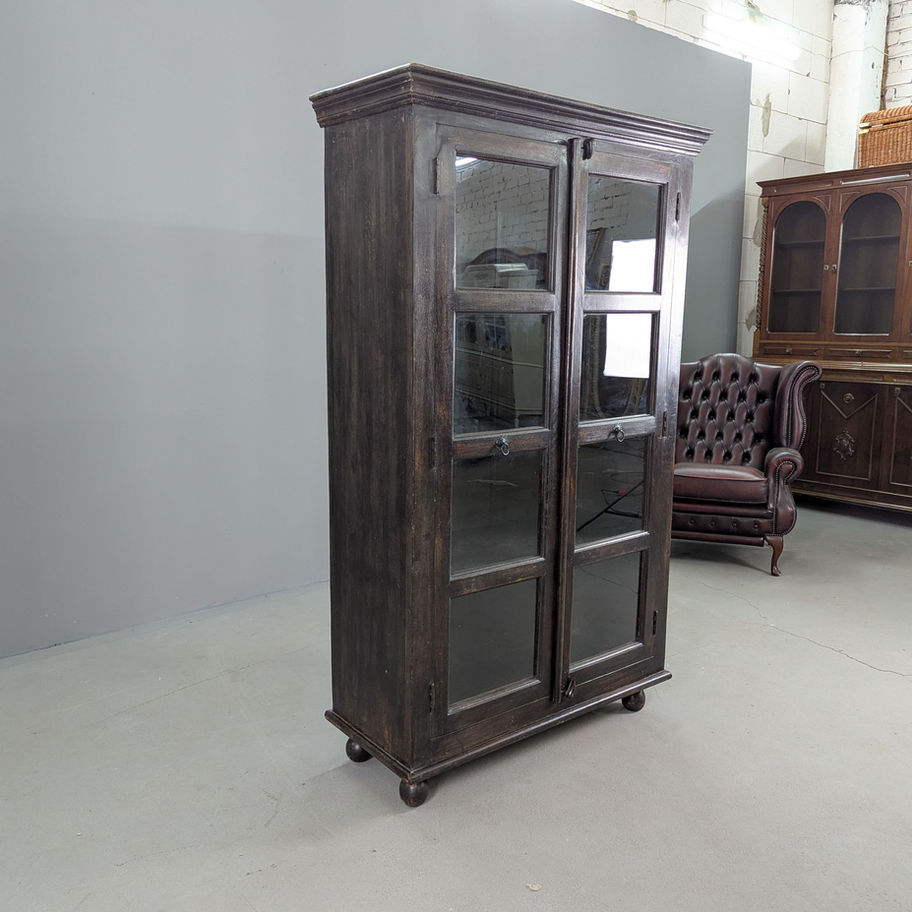 Tamsaus kietmedžio indauja su stiklinėmis durimis, pastatyta prieš pilką foną šalia tamsiai rudo odinio krėslo.