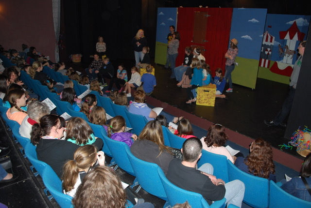 A wide shot of the house of the theater, filled with patrons, and actors on the stage