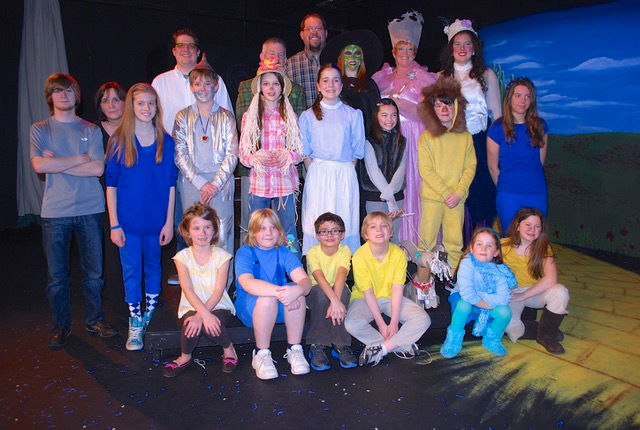 The cast of Wizard of Oz, including Dorothy, the Scarecrow, the Lion, the Tin Man, Glinda, the Wicked Witch of the West, and some munchkins, pose on stage