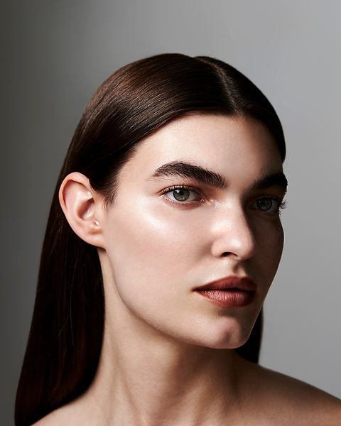 Fashion & Beauty Photography Campaign. Stunning female model in studio with dramatic lighting. Natural makeup with glowing highlighter on cheeks and soft red lips. Full brow and green eyes. Slick shiny hair with centre parting.