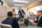 Four corporate workers sitting around a meeting table, two people shaking hands.