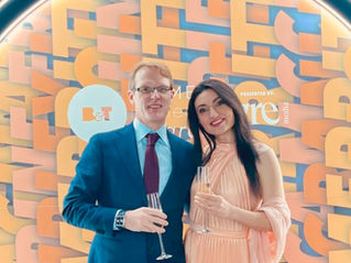 Wayne and Azadeh Williams posing in front of a B&T Media awards orange display, holding champagne glasses.