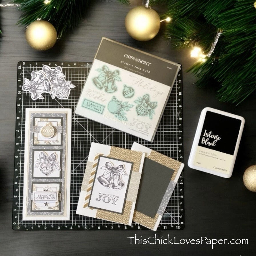 Crafting supplies on a black mat include holiday stamps, cards with "Wishing You Joy," and Intense Black ink. Greenery and gold ornaments in background.