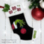Black Christmas stocking with "HO HO HO" and "GRINCH" text. Features green hand holding a red ornament. Red and white decor in background.