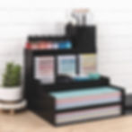 Black desk organizer holds colorful inks, crafting supplies, and paper. Potted plant on the side, set against a white brick wall.