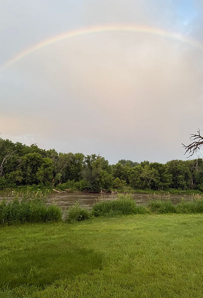 A rainbow above the red river in Moorhead MN wiht a blue sky and green grass and trees
