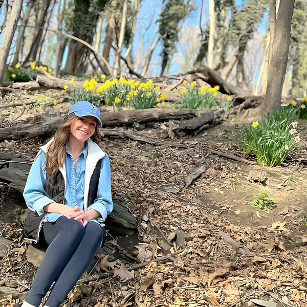 Ashley sitting on the ground smiling near tree roots and daffodils in a wooded area