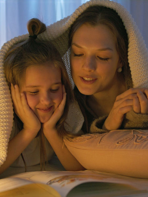 Mother and daughter reading a bedtime book together under a blanket, cozy nighttime routine for kids' emotional connection and literacy development.