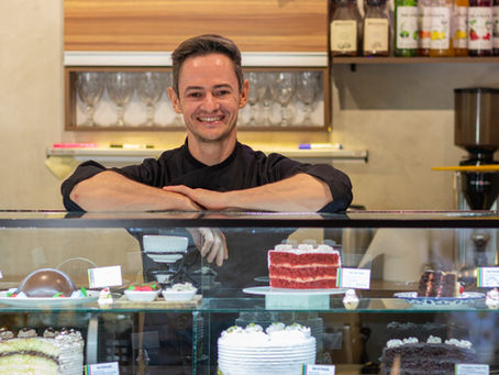 O chef Ronaldo Savaris, proprietário da Cakes Savaris, sorri em frente ao logo de sua confeitaria. Foto por Eduardo Francischelli.