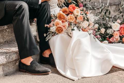 close up photo of bride and groom sitting on steps, the bride is holding a bouquet of peachy coloured roses made by the florist Sass Flower