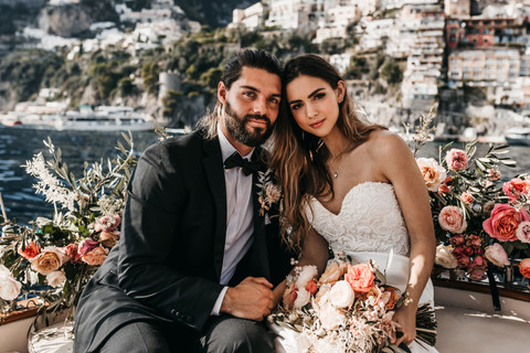bride and groom sitting on a boat surrounded by pink roses with the houses of Positano in the background on their elopement day in the Amalfi Coast