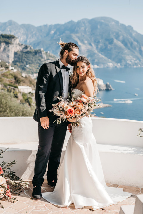 Couple in wedding attire standing under a lemon tree on a terrace overlooking the mountains and sea of the Amalfi coast