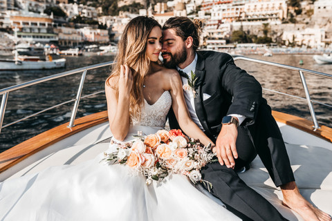 bride and groom sitting on the front of a boat with the houses and mountain side of Positano in the background on their elopement day in the Amalfi Coast