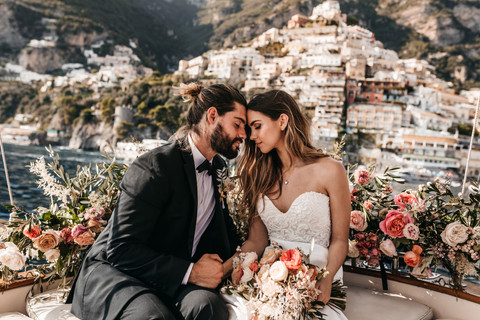 bride and groom sitting on a boat surrounded by pink roses with the houses of Positano in the background on their elopement day in the Amalfi Coast