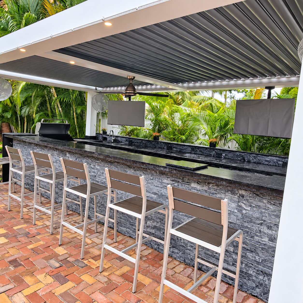 Covered backyard bar with stacked-stone cladding, seating, brick pavers, and tropical landscaping