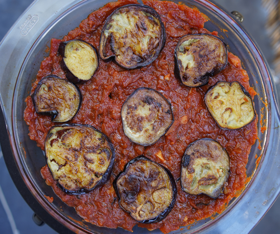 Roasted eggplant slices atop vibrant red tomato sauce in a clear dish. Rich textures and colors, with a cozy, appetizing feel.