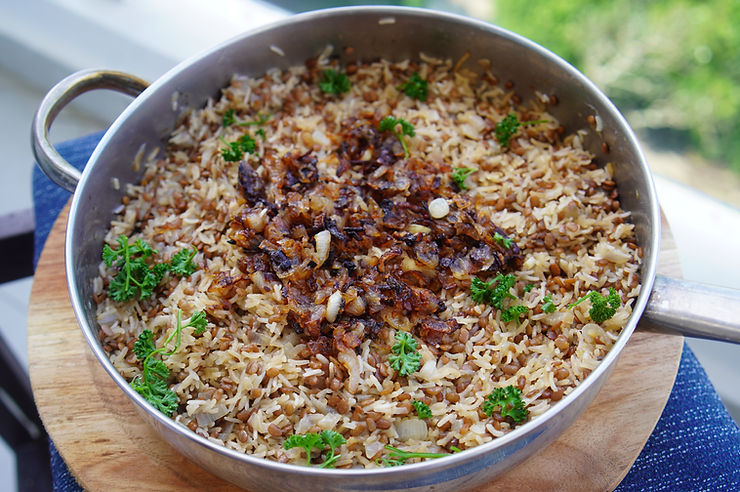 Pan of cooked rice with caramelized onions and green parsley, placed on a wooden board. Bright outdoor setting with greenery in background. Mujaddara