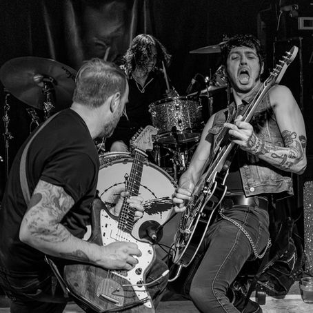 Black-and-white live concert photograph of Torture Twinn performing on stage, featuring two guitarists and drummer in an energetic punk rock performance. The image captures the band's raw live energy, intense stage presence, and underground punk atmosphere during a club show.
