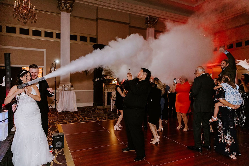 Bride in white gown fires a smoke cannon at a wedding reception as guests dance and cheer in a grand hall with red lighting.