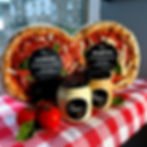 Bangin’ Pizza products displayed on a checkered table: two wrapped stone-baked pizzas with black labels and three branded sauce jars, arranged alongside fresh tomatoes and basil near a bright window.
