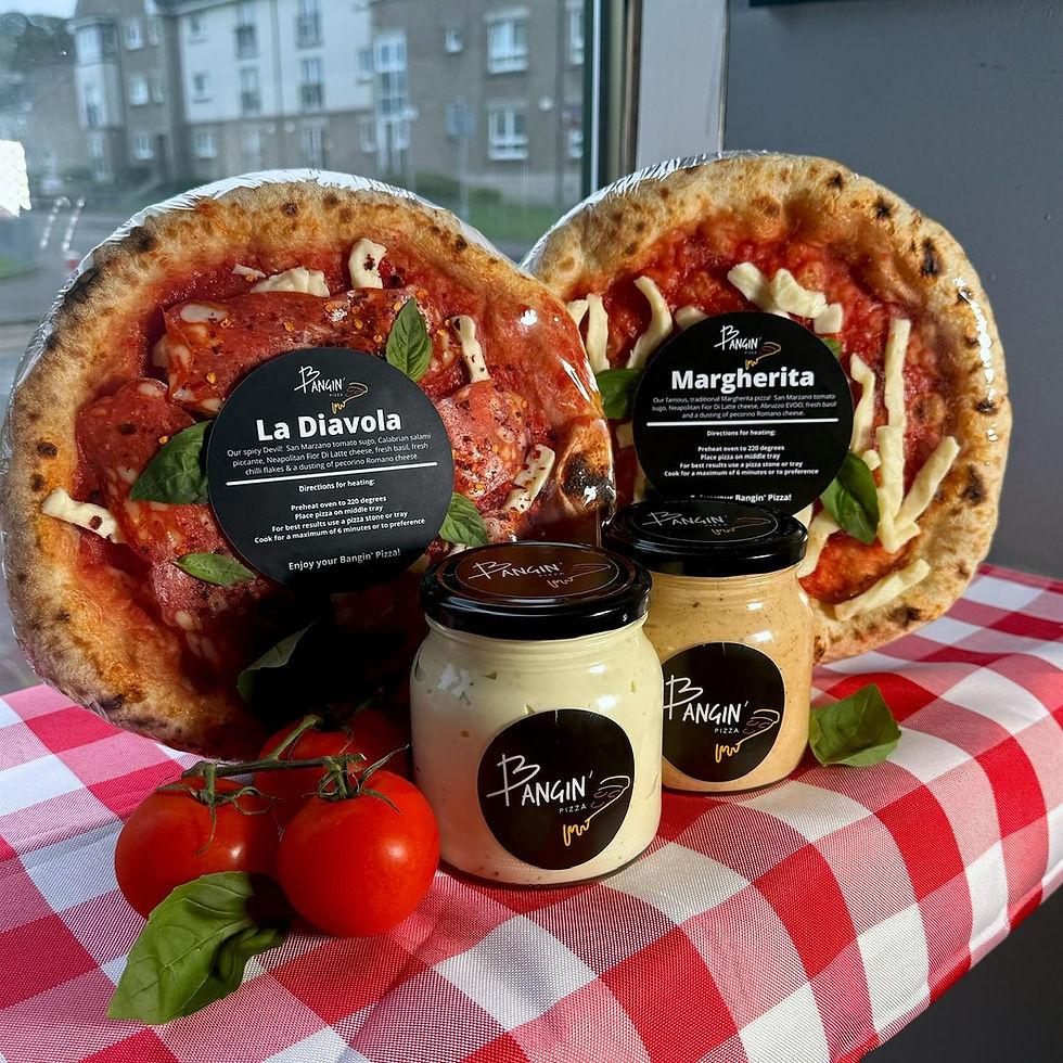 Two Bangin’ Pizza part baked pizzas, La Diavola and Margherita, standing upright on a red-and-white checkered tablecloth beside three Bangin’ Pizza dipping sauce jars. Fresh tomatoes and basil sit in front, with a window and residential buildings of Dumbarton blurred in the background.