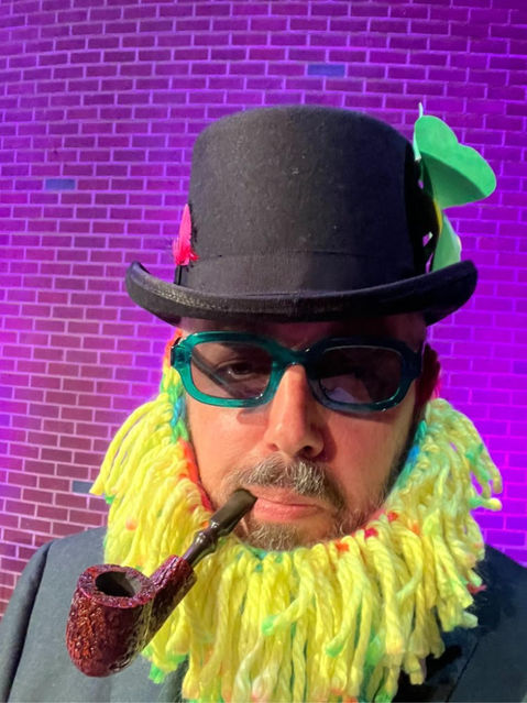 Sean with black hat, yellow ruff, green sunglasses, pipe in moth, purple background with brick pattern