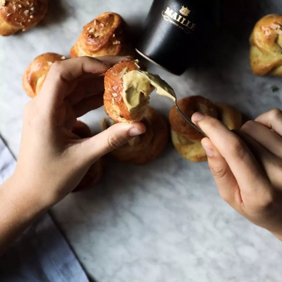 hands apply a spoon full of mustard to a piece of bread knot. in the background are more bread knots on a marble table and a black jar of mustard