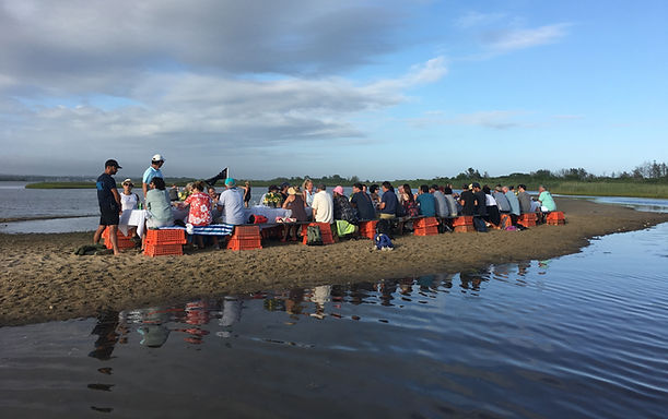 A group of people sitting on lobster crates at a makeshift table on a sandbar