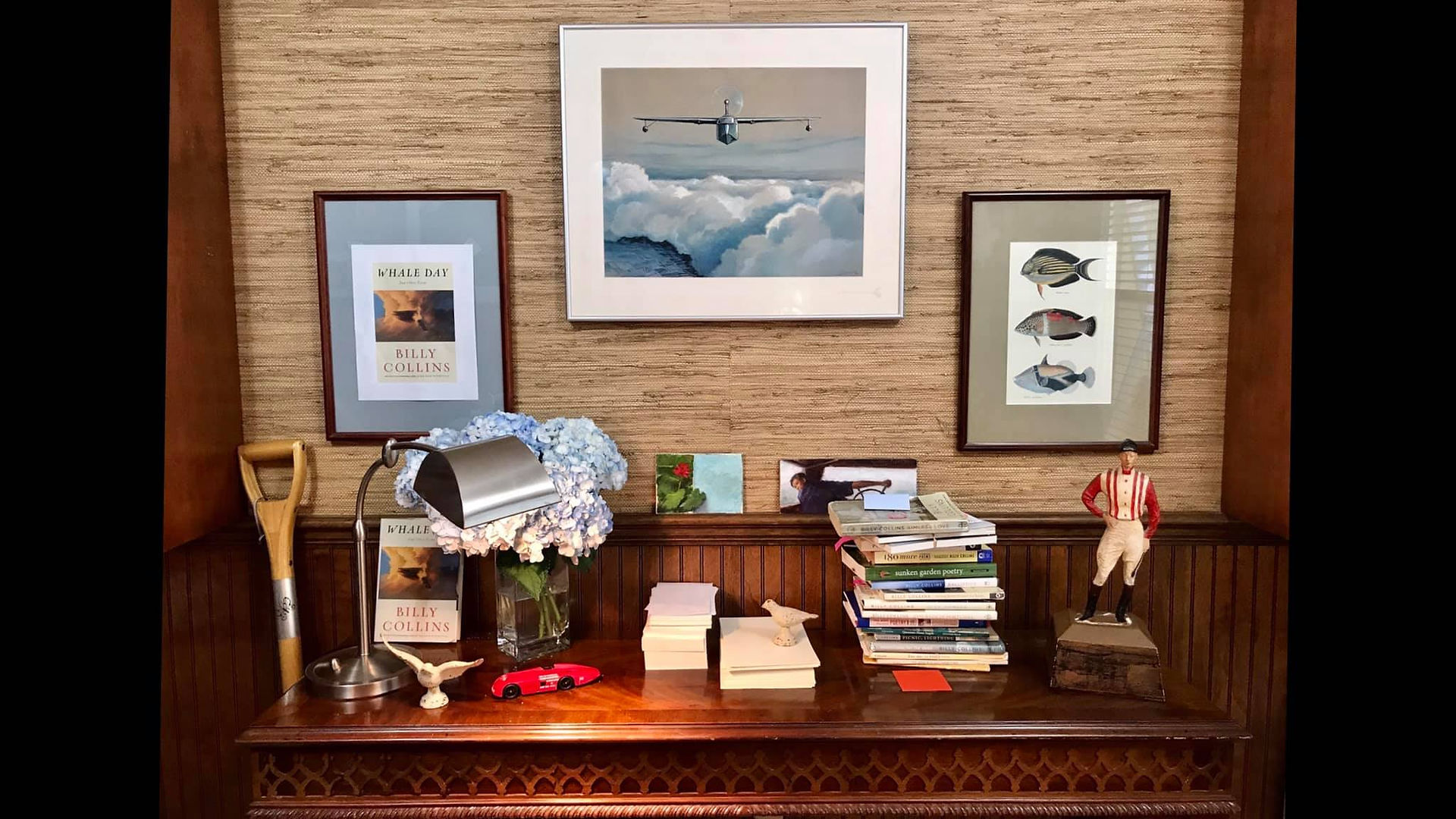 a table, covered in books and small trinkets, in front of a wall with beige grasscloth wallpaper, natural wood wainscoting, and three framed pictures
