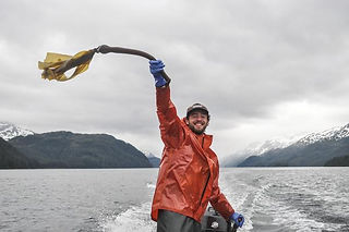 a smiling man in a red raincoat sitting on a boat on a grey hazy ocean waves a piece of kelp over his head