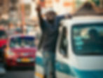 Ghana trotro mate leans out of a white and blue van gesturing, with busy cars and blurred background. Vibrant colors suggest a lively urban scene.