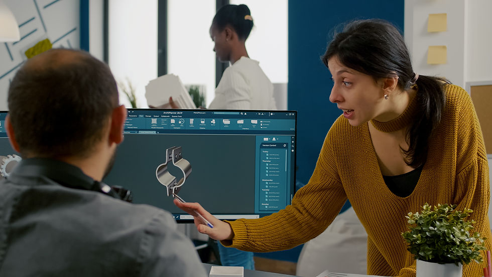 Woman in mustard sweater points at computer screen displaying 3D design. Man listens. Office setting, another person in background.