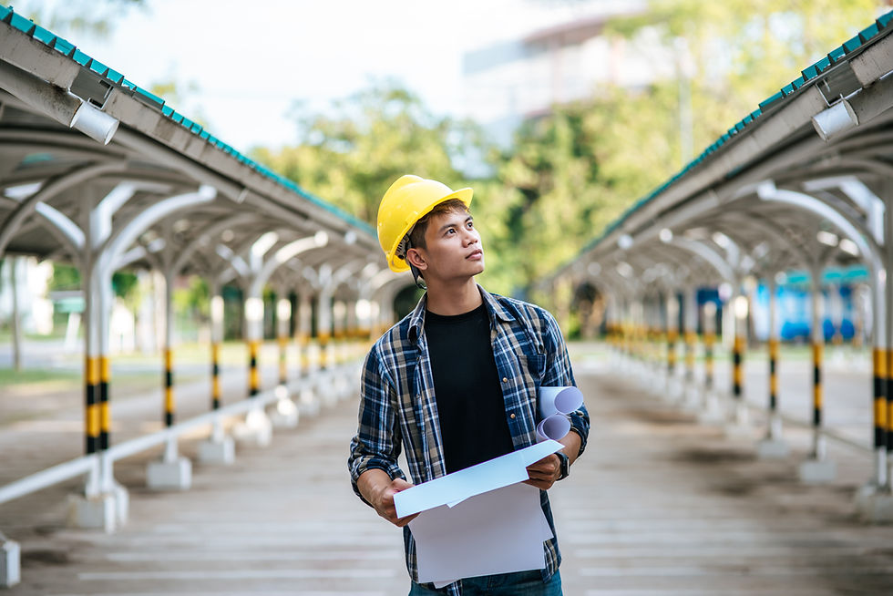 Young man in a yellow hard hat holds blueprints, looking upwards. He's on a paved path between metal structures, with greenery in the background.